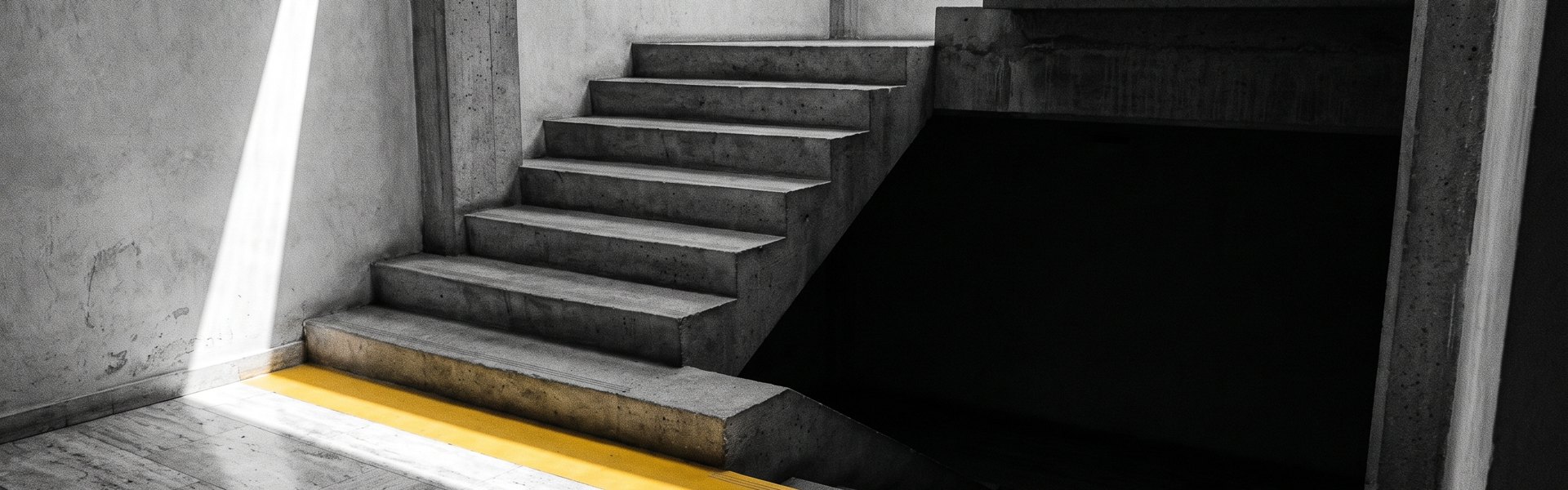 Monolithic concrete stairwell, sun blade on travertine floor, chrome-yellow line disappearing into shadow. Black and white -- Creatopy alternatives.