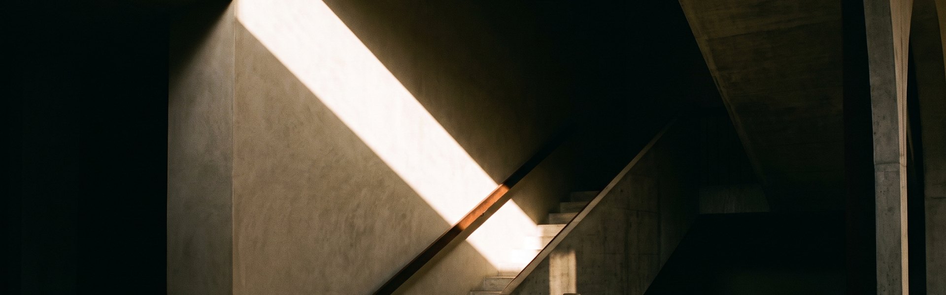 A single blade of sunlight illuminates a section of a concrete stairwell ascending alongside a lime plaster wall into shadow -- AI Creative Maturity Model.